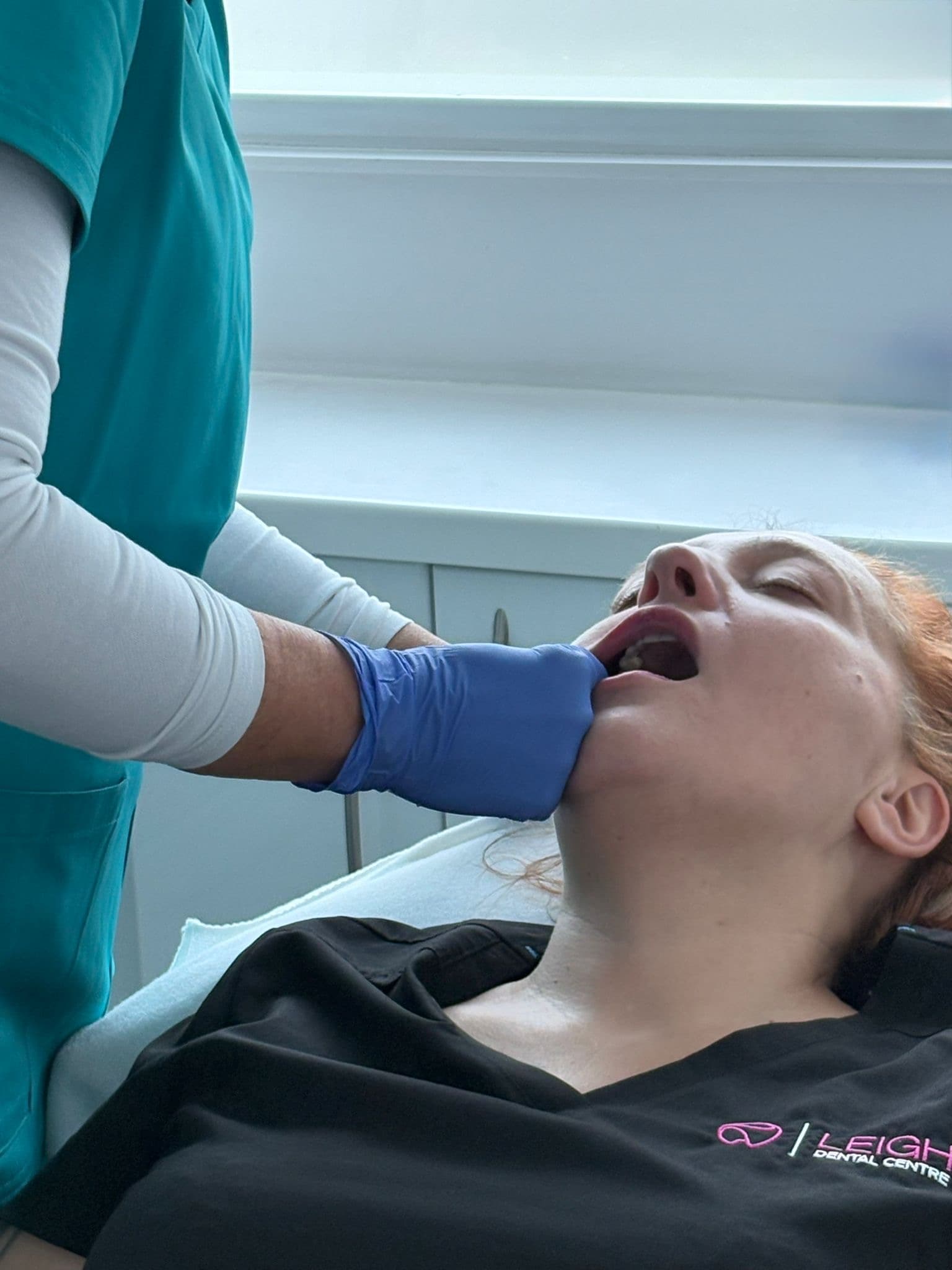 Dentist wearing blue gloves examines a patient's open mouth during a clinical procedure.