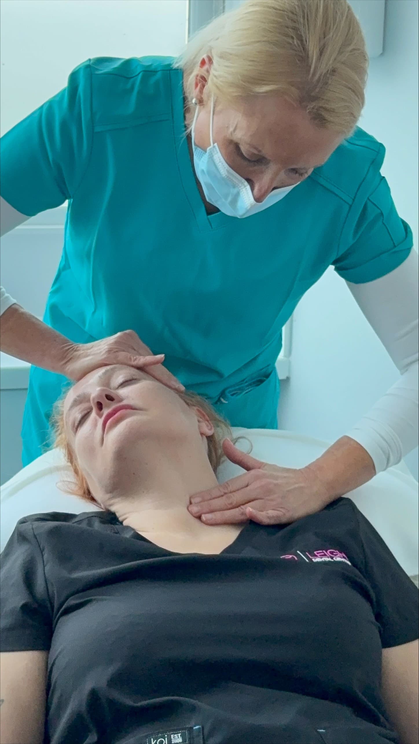 Medical professional in teal scrubs performing a neck and head examination on a reclining patient.