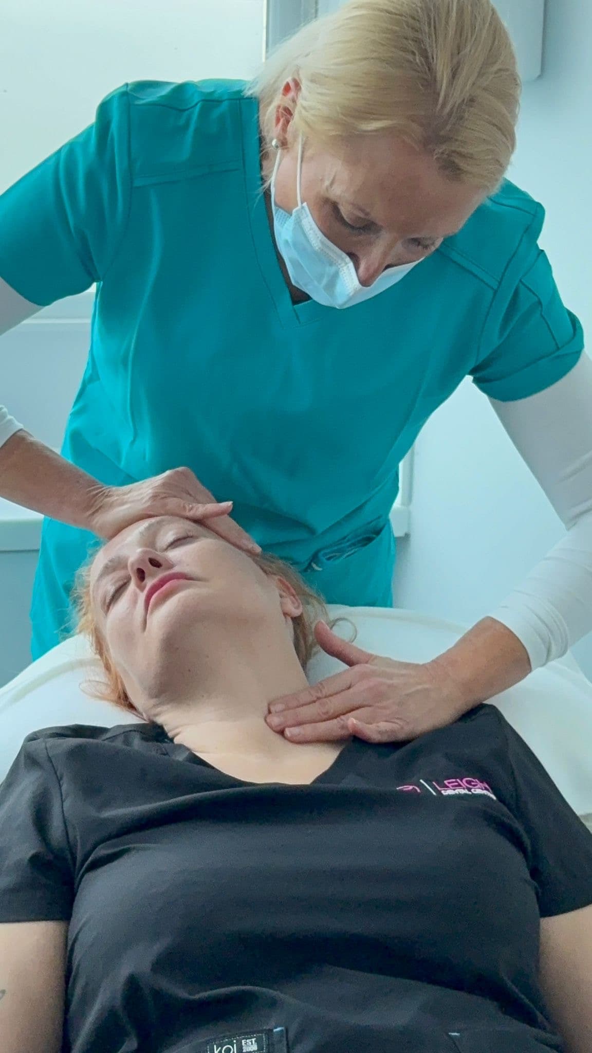 Medical professional in teal scrubs performing a neck and head massage on a reclining patient.