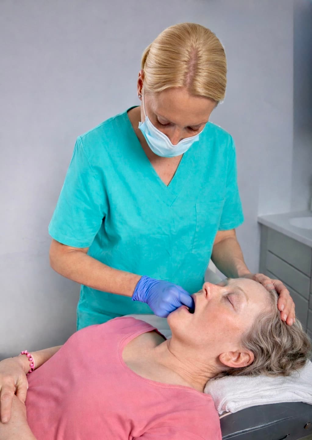 Masked healthcare professional in teal scrubs performing an intraoral manual therapy treatment on a patient.